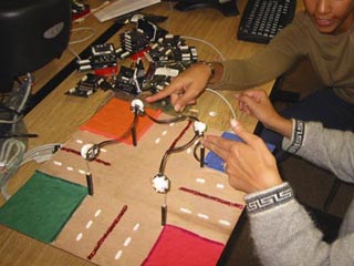 A photo of two students working on what looks like a minature four-way intersection. A photo of two students working on what looks like a minature four-way intersection.