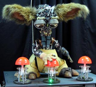 A photograph of the robot Leo. Leo stands before three plunger style buttons with its hand on the center button. A photograph of the robot Leo. Leo stands before three plunger style buttons with its hand on the center button.