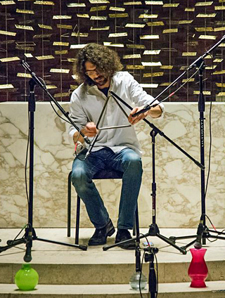 Photo of a seated man striking a metal triangle, with microphones. Photo of a seated man striking a metal triangle, with microphones.