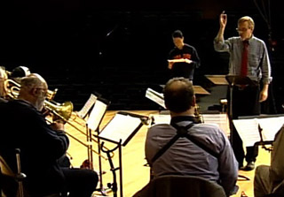 Photo of part of a large jazz ensemble on stage - saxophones and trombones - with conductor and composer out front. Photo of part of a large jazz ensemble on stage - saxophones and trombones - with conductor and composer out front.