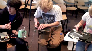 Still photo from video of class activity, showing three students working on a sonic circuit. Still photo from video of class activity, showing three students working on a sonic circuit.