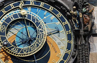 Photo of an ornate clockface with sun and moon hands, adjacent to statues of a skeleton and musician. Photo of an ornate clockface with sun and moon hands, adjacent to statues of a skeleton and musician.