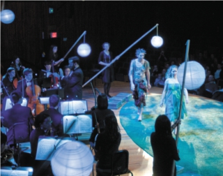 Puck pursues Peaseblossom across a clearing, surrounded by large paper lanterns on tall bamboo poles. An orchestra plays behind them. Puck pursues Peaseblossom across a clearing, surrounded by large paper lanterns on tall bamboo poles. An orchestra plays behind them.