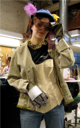 Over street clothes, a woman wears a corset mock-up, a jacket with painted Celtic knots, gauntlets with metal panels and wires, a gold-and-silver bead necklace, a purple-and-gold mask, and a blue hat with a yellow lily. Over street clothes, a woman wears a corset mock-up, a jacket with painted Celtic knots, gauntlets with metal panels and wires, a gold-and-silver bead necklace, a purple-and-gold mask, and a blue hat with a yellow lily.