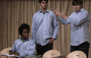 One student sits at a desk, while two others stand beside him, gesturing. One student sits at a desk, while two others stand beside him, gesturing.