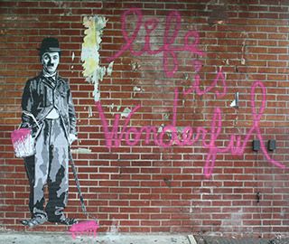 The surface of a red-brick building shows an illustration of a man with a small moustache, tight suit coat, and baggy pants, holding a paint can and roller. He stands next to the words "life is wonderful" painted in hot pink paint. The surface of a red-brick building shows an illustration of a man with a small moustache, tight suit coat, and baggy pants, holding a paint can and roller. He stands next to the words "life is wonderful" painted in hot pink paint.