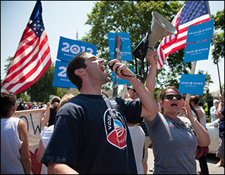 A photograph of two campaign field organizers, wearing Obama 2012 shirts. One is shouting into a megaphone and the other is cheering. The crowd behind them is holding many Obama / Biden 2012 signs and American flags. A photograph of two campaign field organizers, wearing Obama 2012 shirts. One is shouting into a megaphone and the other is cheering. The crowd behind them is holding many Obama / Biden 2012 signs and American flags.