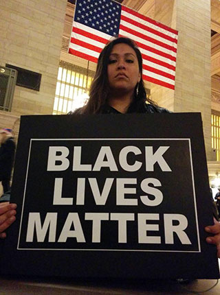 A solemn-faced brown-skinned woman, seated beneath the American flag, holds a sign with the text: Black Lives Matter. A solemn-faced brown-skinned woman, seated beneath the American flag, holds a sign with the text: Black Lives Matter.