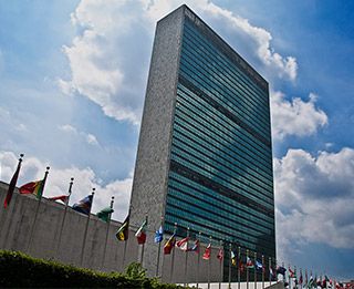 A tall glass and concrete structure fronted by the flags of various nations stands against a blue sky. A tall glass and concrete structure fronted by the flags of various nations stands against a blue sky.