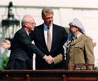Two gray-haired gentleman, one wearing a suit, the other wearing a brown jacket and a long scarf, shake hands while a younger man stands between them. Two gray-haired gentleman, one wearing a suit, the other wearing a brown jacket and a long scarf, shake hands while a younger man stands between them.
