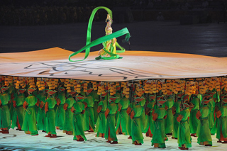 Dozens of people dressed in bright green uniforms carry poles that support a movable stage. A dancer twirling a green ribbon, stands on the stage. Dozens of people dressed in bright green uniforms carry poles that support a movable stage. A dancer twirling a green ribbon, stands on the stage.
