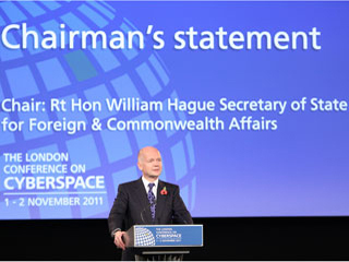 A slim, formally dressed balding man stands at a podium in front of a huge projection screen. A slim, formally dressed balding man stands at a podium in front of a huge projection screen.