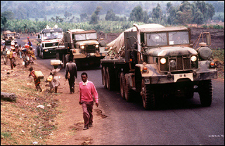 Three military vehicles pass alongside walking villagers on a narrow road. Three military vehicles pass alongside walking villagers on a narrow road.