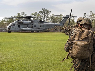 A photograph of a line of Marines wearing camouflage walking across a grass field to board a large helicopter. A photograph of a line of Marines wearing camouflage walking across a grass field to board a large helicopter.