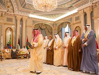 In the middle of a wide-open, elaborate room, six men covered in fabric from head to toe stand behind a similarly-garbed gentleman who holds a binder. In the middle of a wide-open, elaborate room, six men covered in fabric from head to toe stand behind a similarly-garbed gentleman who holds a binder.