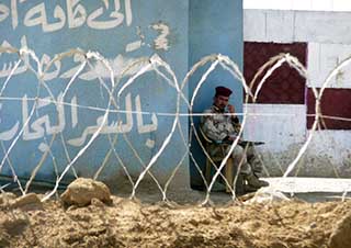 A photo of an Iraqi Army soldier seated behind a barbed wire fence. A photo of an Iraqi Army soldier seated behind a barbed wire fence.