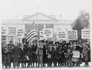 Wives of political prisoners standing in a crowd with protest signs and an American flag. Wives of political prisoners standing in a crowd with protest signs and an American flag.