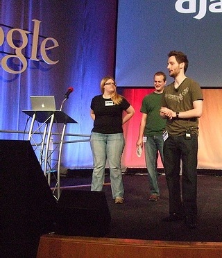 A photo of three people on a presentation stage at a conference. A photo of three people on a presentation stage at a conference.