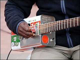 A picture of a man playing an electric guitar. The guitar’s body is made out of an old Castrol oil can, which has been painted with South African colors (black, green, and red). A picture of a man playing an electric guitar. The guitar’s body is made out of an old Castrol oil can, which has been painted with South African colors (black, green, and red).