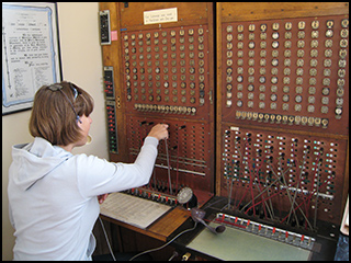 A photograph of a woman plugging a cable into a slot on an old telephone switchboard. A photograph of a woman plugging a cable into a slot on an old telephone switchboard.