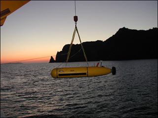 A small submarine-type vehicle being lowered into the water from a large ship at sunset near a rocky coast. A small submarine-type vehicle being lowered into the water from a large ship at sunset near a rocky coast.
