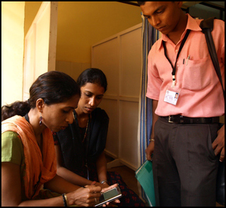 A female health worker logs data on a device as two others look on. A female health worker logs data on a device as two others look on.