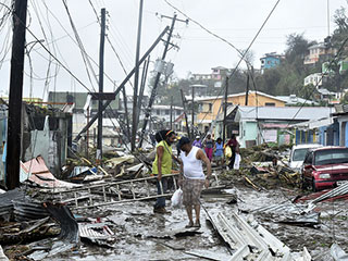 A photo of two men standing in debris after Hurricane Maria hit Puerto Rico. They are surrounded by damaged houses and fallen telephone poles and wires. A photo of two men standing in debris after Hurricane Maria hit Puerto Rico. They are surrounded by damaged houses and fallen telephone poles and wires.