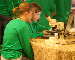 Two girls in green shirts use a laptop computer and microscope. Two girls in green shirts use a laptop computer and microscope.