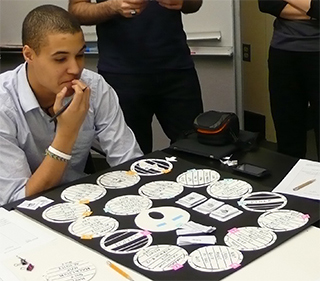 A student is seated at a table, looking down at a homemade board game. The game consists of a series of round spaces that the player must move through. A student is seated at a table, looking down at a homemade board game. The game consists of a series of round spaces that the player must move through.