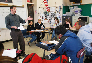 A teacher stands in a classroom with students seated and taking notes. The governor of Massachusetts sits with the students. A teacher stands in a classroom with students seated and taking notes. The governor of Massachusetts sits with the students.
