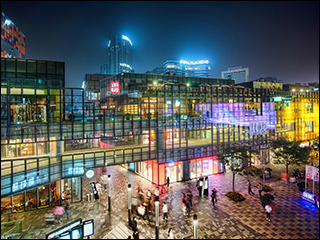 A photograph of a city at night time. There is a plaza in the foreground, a large glass building in the middle, and a city skyline in the background. Many of the buildings have neon lights of all colors. The lights are reflected off the glass building. A photograph of a city at night time. There is a plaza in the foreground, a large glass building in the middle, and a city skyline in the background. Many of the buildings have neon lights of all colors. The lights are reflected off the glass building.