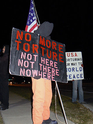 A protester stands with a black hood over his face while holding a sign that reads “No more torture, not here, not there, nowhere.” A protester stands with a black hood over his face while holding a sign that reads “No more torture, not here, not there, nowhere.”