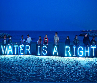 Protesters stand on a beach at nightfall with a neon blue sign that reads “Water is a right”. Protesters stand on a beach at nightfall with a neon blue sign that reads “Water is a right”.