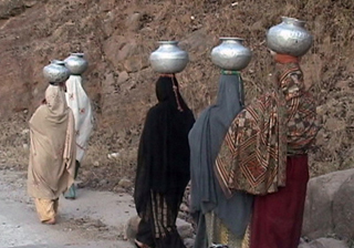 Pakistani women clad in traditional clothing carrying water in metalic vessels on their heads . Pakistani women clad in traditional clothing carrying water in metalic vessels on their heads .