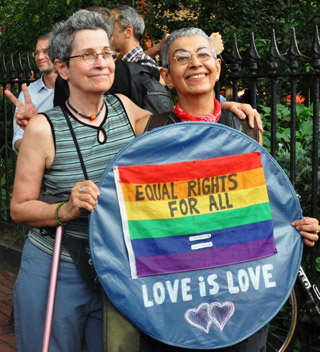 A middle-aged, grey-haired female couple pose with their arms around each other,holding a sign with the words: Equal Rights For All. Love Is Love. A middle-aged, grey-haired female couple pose with their arms around each other,holding a sign with the words: Equal Rights For All. Love Is Love.