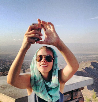 A photograph of a woman holding a cell phone in front of her taking a picture of herself against a landscape of mountains. A photograph of a woman holding a cell phone in front of her taking a picture of herself against a landscape of mountains.
