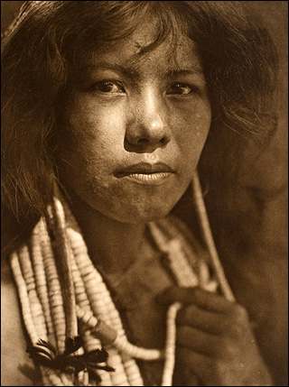 A sepia-toned photograph of a Native American young woman. She has beaded necklaces wrapped around her neck and is looking directly into the camera. A sepia-toned photograph of a Native American young woman. She has beaded necklaces wrapped around her neck and is looking directly into the camera.