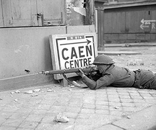 A European soldier lies on his stomach aiming a rifle. There is rubble around him and a sign pointing towards Caen Centre. A European soldier lies on his stomach aiming a rifle. There is rubble around him and a sign pointing towards Caen Centre.