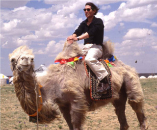 Photo of Dr. Wheatley riding a camel in front of a row of white tents. Photo of Dr. Wheatley riding a camel in front of a row of white tents.