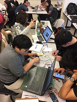 Several students sitting around a rectangular table. Theyre manipulating electronics.
