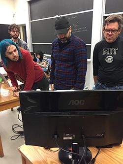 Three men, in a classroom setting, looking a computer monitor.
