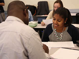 Photo of part of a classroom where 2 students are talking and more students behind them. Photo of part of a classroom where 2 students are talking and more students behind them.